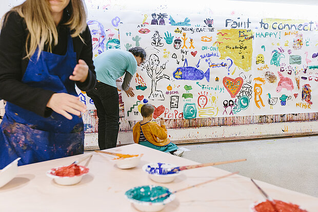 
            
                auf einem Tisch stehen Farbtöpfe, eine Person arbeitet mit den Farben, im Hintergrund eine weiße Wand voller bunter Zeichnungen, eine Frau scheint einem Kind dort etwas zu zeigen
            
        