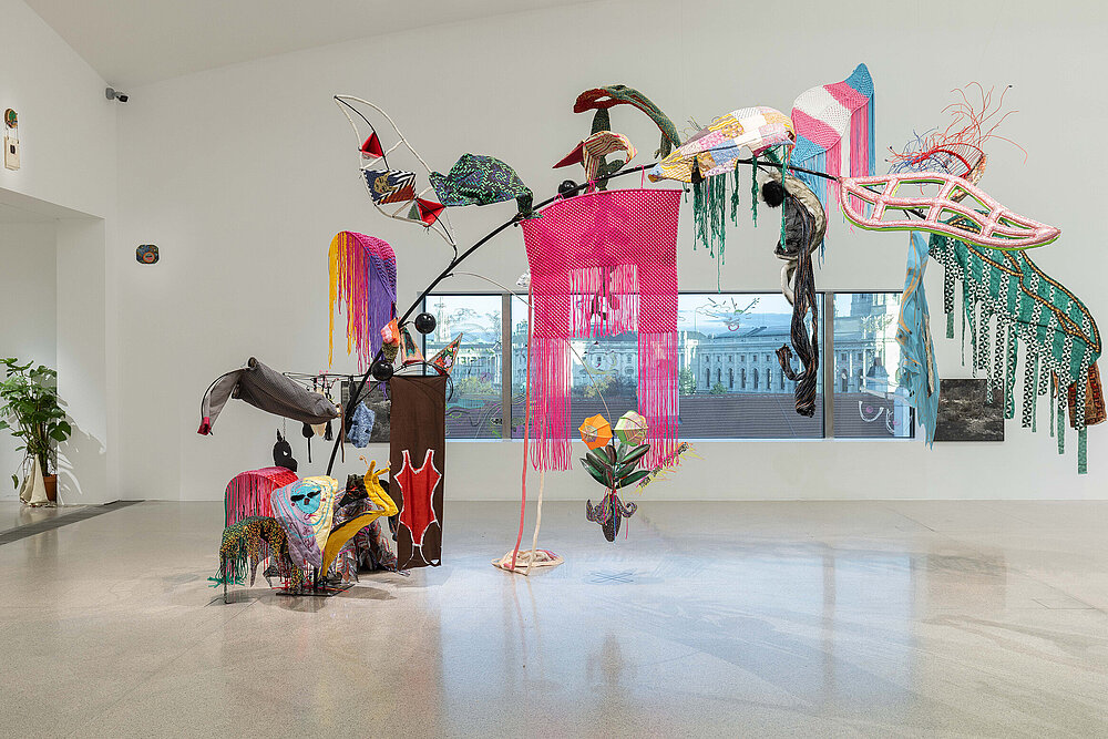  A large-format sculpture is placed in the exhibition space in front of an elongated window. Various coloured fabrics and structures hang down from it. 