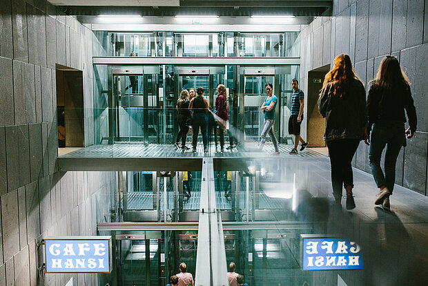 
            
                Blick ins mumok Treppenhaus auf gläserne Aufzüge, Menschen stehen vor den Aufzügen und gehen auf sie zu
            
        