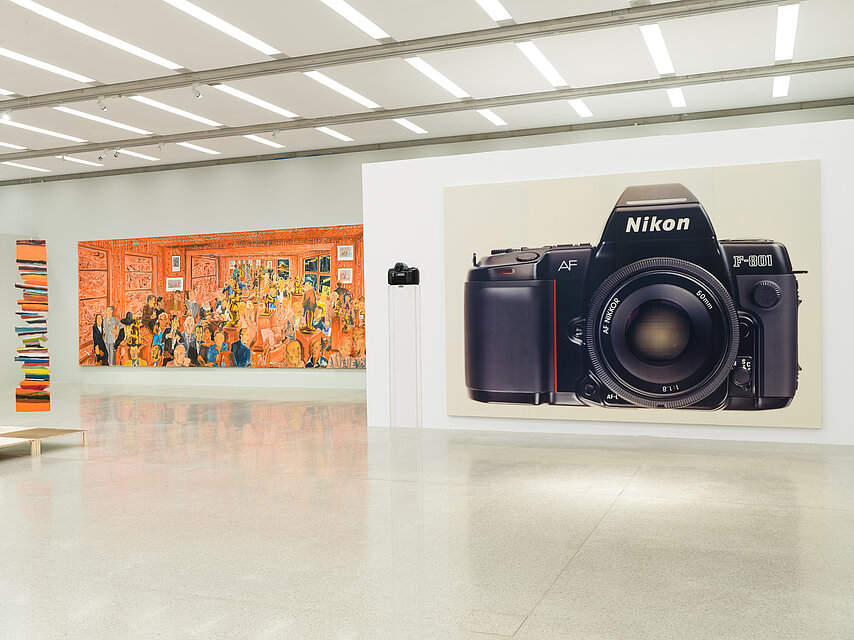 Exhibition room with white walls and light floor, on the left in the background an orange, colourful painting with many people, on the right a large picture of a camera
