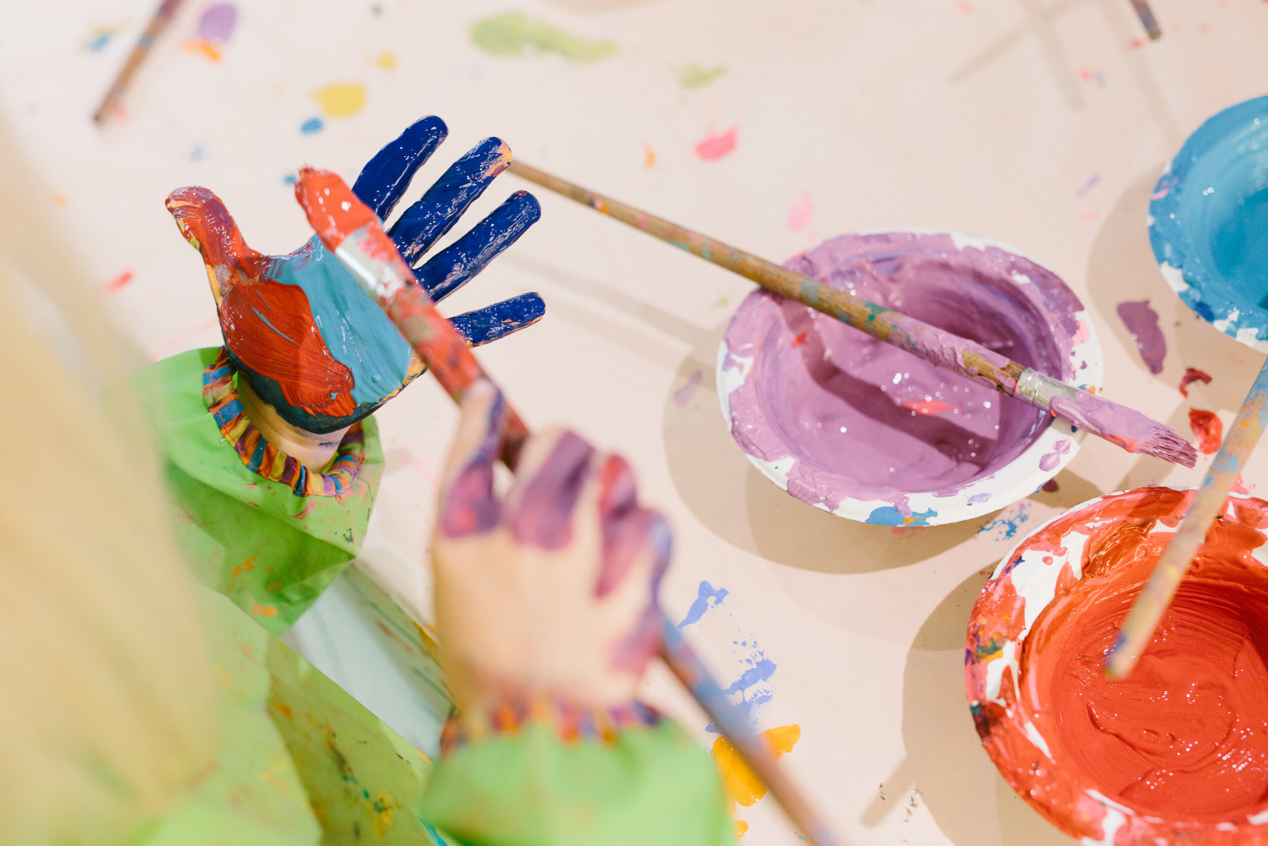 Kinderhände spielen mit Farben und Pinseln, die Hand ist bunt bemalt, auf dem Tisch stehen Farbtöpfe