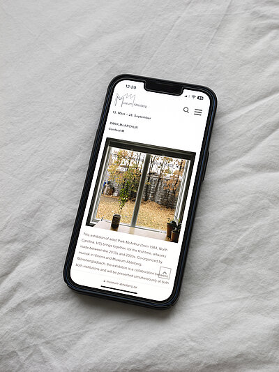  A close up of a smartphone displaying Museum Abteiberg’s webpage for the exhibition Contact M by Park McArthur. The phone rests on a white comforter.