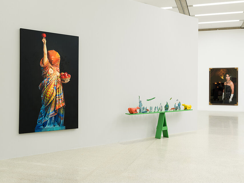 Works of art in a bright exhibition space, on the left a woman posing wrapped in colourful scarves like the Statue of Liberty, painted on a black background