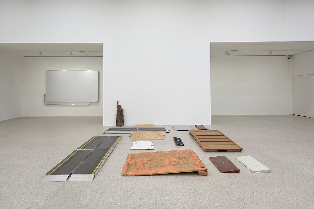 A collection of wooden and metal ramps arranged on the gallery’s floor in a structured yet fragmented manner. The materials show signs of wear, with some featuring chipped paint and rust. A wall-mounted artwork composed of aluminum panels made from Austrian highway signs, blank yet retaining their original format, is mounted to an alcove wall in the background.