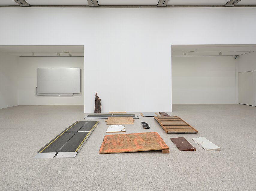 A collection of wooden and metal ramps arranged on the gallery’s floor in a structured yet fragmented manner. The materials show signs of wear, with some featuring chipped paint and rust. A wall-mounted artwork composed of aluminum panels made from Austrian highway signs, blank yet retaining their original format, is mounted to an alcove wall in the background.