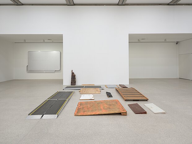 
            
                A collection of wooden and metal ramps arranged on the gallery’s floor in a structured yet fragmented manner. The materials show signs of wear, with some featuring chipped paint and rust. A wall-mounted artwork composed of aluminum panels made from Austrian highway signs, blank yet retaining their original format, is mounted to an alcove wall in the background.
            
        