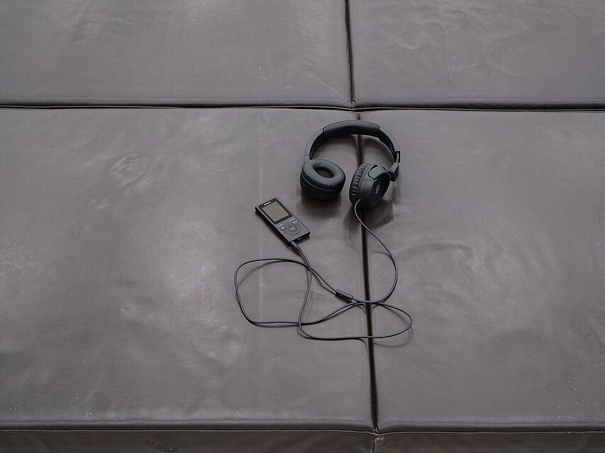 A pair of headphones connected to a portable audio device lie on a grid of dark leather bench cushions.