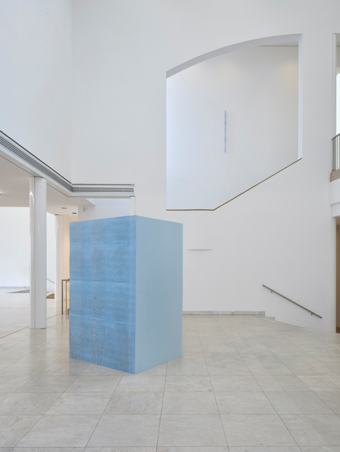 A freestanding rectangular sculpture made of blue polyurethane foam. In the background, high on a wall above a set of stairs, a sculpture of stacked ventilator filters hangs. The filter sculpture is visually framed by a design element: a cut out in the museum wall. The gallery space has high ceilings, arched architectural elements, and staircases leading to different levels.