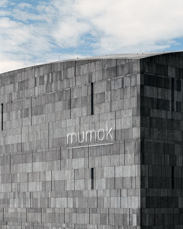 
            
                Fassade des mumok, ein schlichtes Gebäude aus grauem Basaltlavastein mit dem Logo des mumok, im Hintergrund blauer Himmel mit wenigen Wolken
            
        
