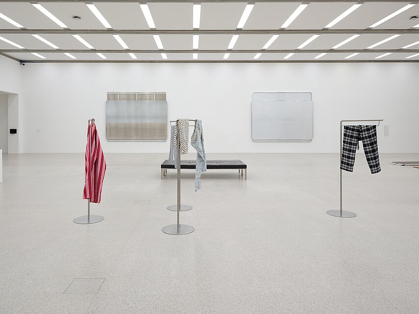 A wide gallery view of multiple artworks, including two large sculptures made of unadorned American and Austrian highway signs. These sculptures resemble one another in their dimension, composition, and softly reflective aluminum surfaces. To the left, a rubber loading dock bumper is bolted to the gallery wall. In the foreground sit two dark leather museum benches and four sculptures made of tall metal stands from which torn pajama pants of various patterns, colors, and fabrics hang.