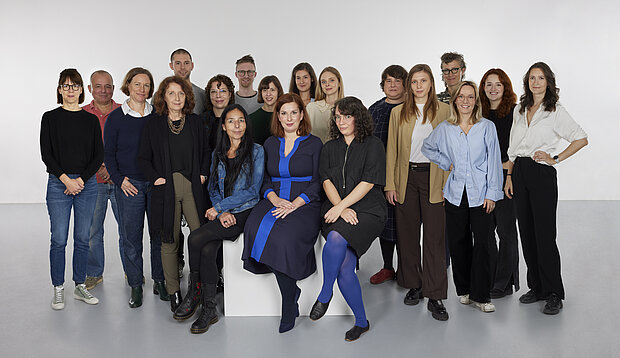 
            
                a group of people in front of a white wall, three of them sitting on a pedestal in the foreground, the team collection at mumok
            
        