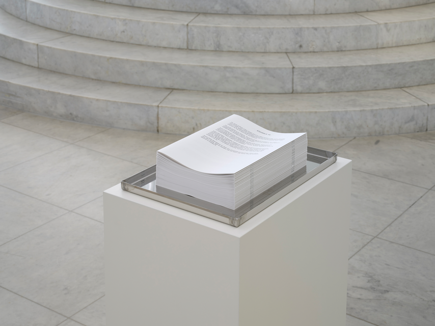 A tall stack of letter-sized pamphlets offered inside of a shallow stainless steel tray. The tray rests on a white pedestal in a gallery space with a curved marble staircase in the background.