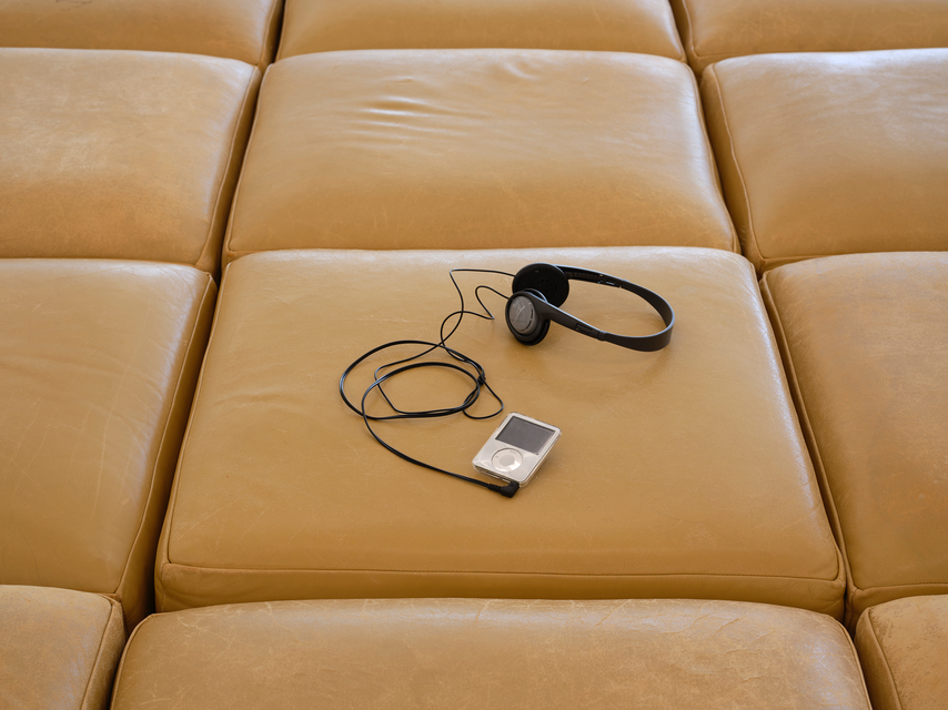 A pair of headphones connected to an iPod lie on a grid-like arrangement of large, square leather cushions that are tan in color. The cushions have visible creases and signs of wear.