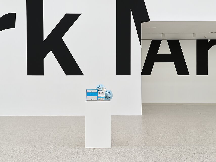 In the foreground sits a white pedestal holding a stainless steel tray filled with barrier creams, a pair of blue foam heel protectors, and individually packaged bandages. In the background, large black text partially spelling "k MAr" stretches across a white wall into an alcove leading to another section of the gallery.