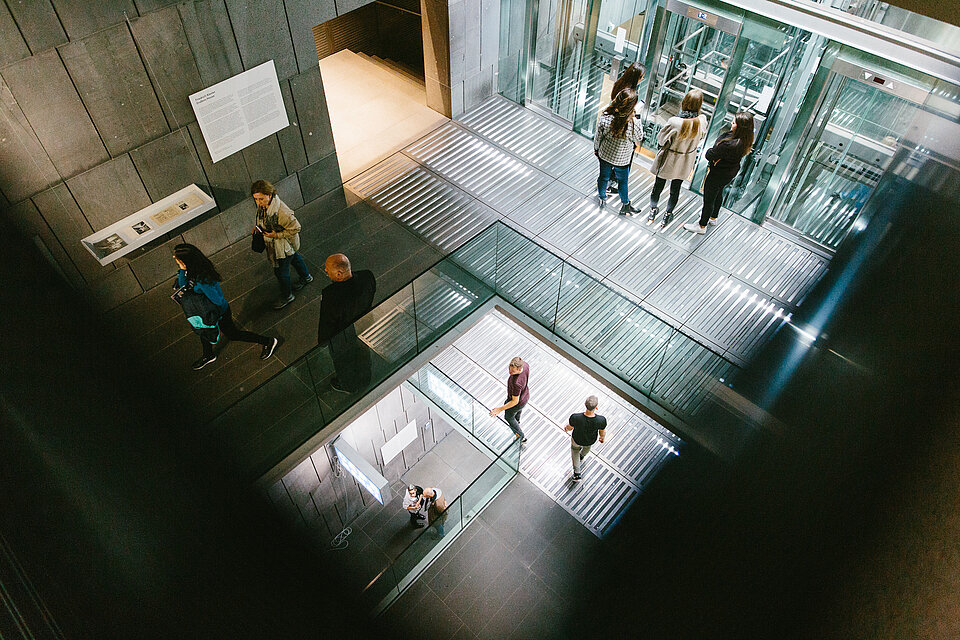 
            
                Blick ins mumok Treppenhaus, gläserne Lifte und Geländer, Wände aus dunklem Stein, verschiedene Menschen stehen auf unterschiedlichen Ebenen im Treppenhaus
            
        