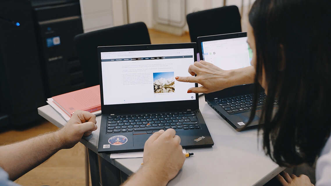 auf einem Tisch stehen zwei Laptops, man sieht von links zwei Hände, die am Laptop arbeiten, von rechts zeigt eine Frau auf den Bildschirm