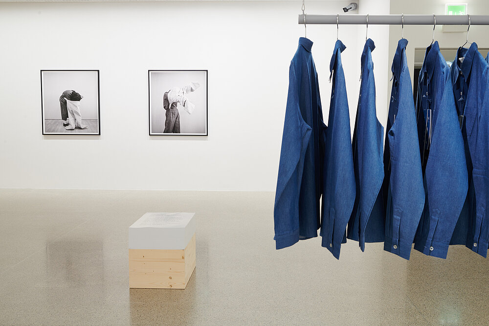 blue shirts on a clothes rail on the right, black and white photographs of people in the background on the left, a wooden box in front