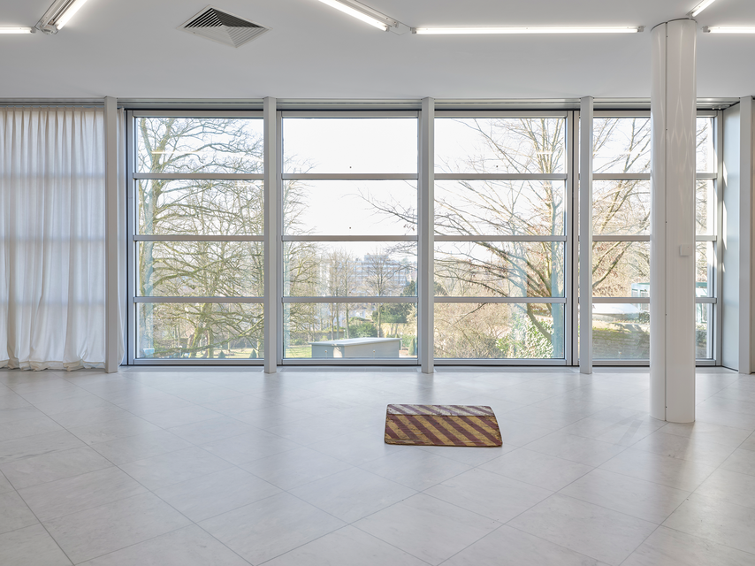 A small ramp with a yellow and red striped pattern, placed directly on the gallery floor. Positioned in front of large windows, the ramp contrasts with the surrounding architecture. Natural light fills the room, with a backdrop of trees and a cityscape visible through the glass